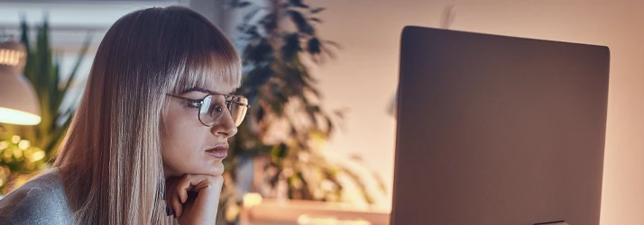 Person wearing eyeglasses looking at a computer monitor while resting their chin on a hand in a dimly lit indoor setting.